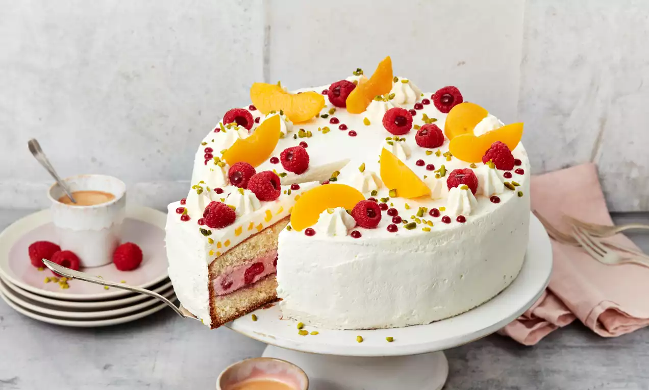 Bijela torta s glatkom kremom, ukrašena kriškama breskve, malinama, šlagom i usitnjenim pistacijama, poslužena na bijelom stalku