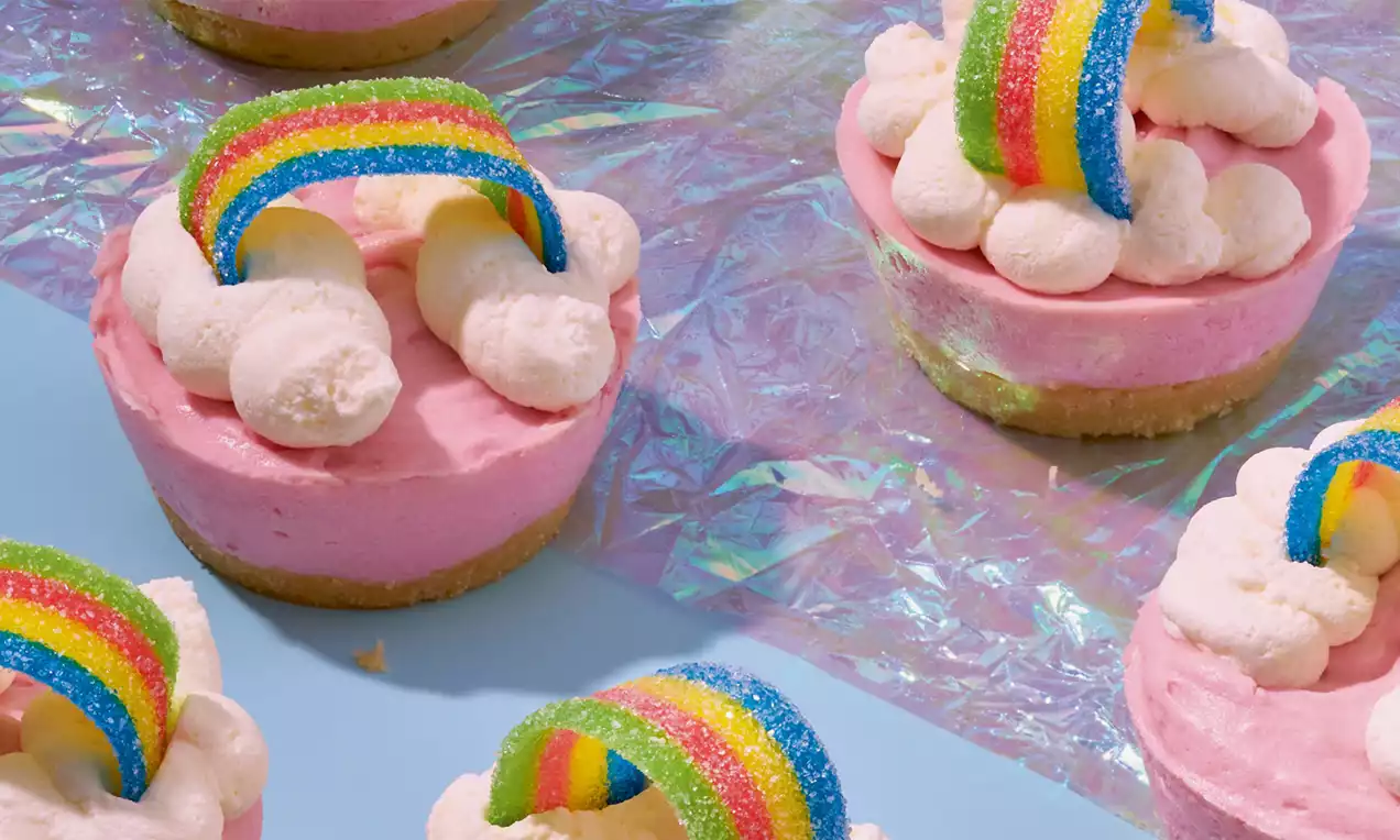 Roze mini-kwarktaartjes met een koekbodem, witte slagroomwolken en kleurrijke regenboogdecoratie, gepresenteerd op glanzend holografisch folie