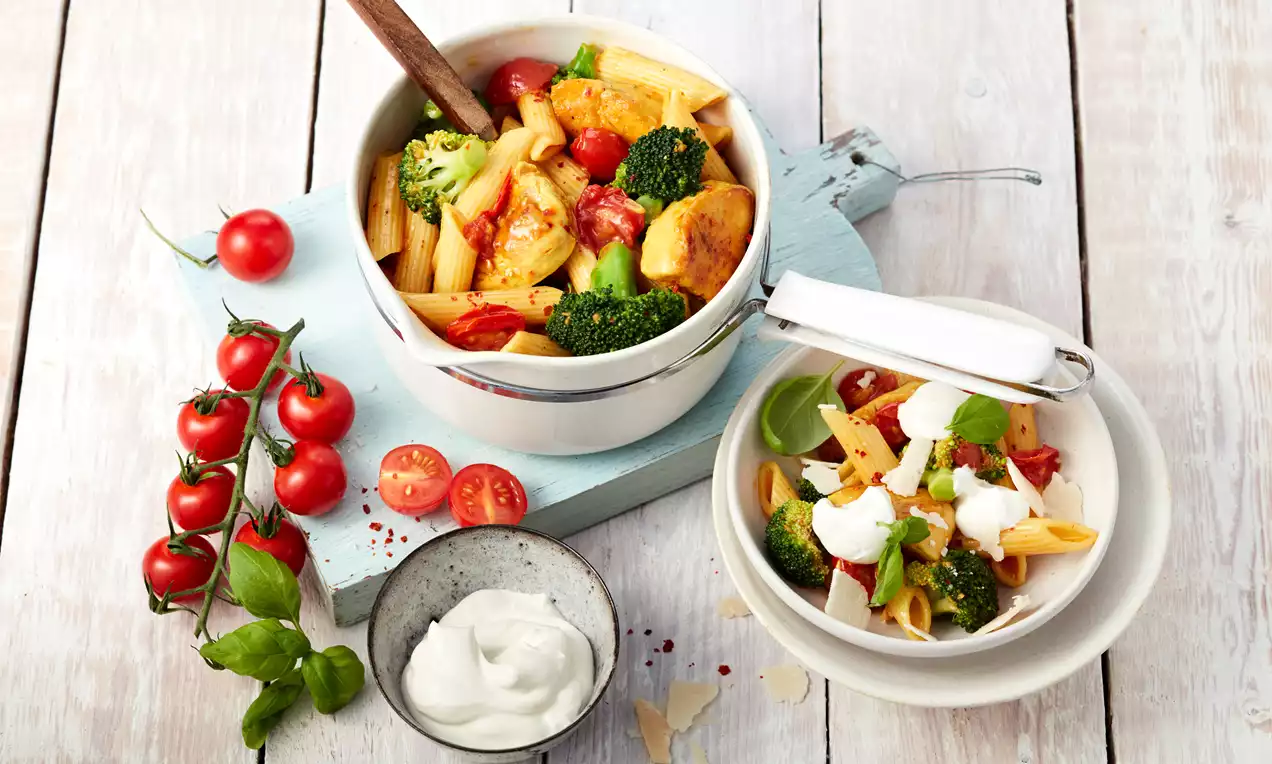 Bunte One-Pot-Pasta mit goldbraunem Hähnchen, knackigem Brokkoli und roten Tomaten in weißem Topf und auf Teller mit frischen Beilagen