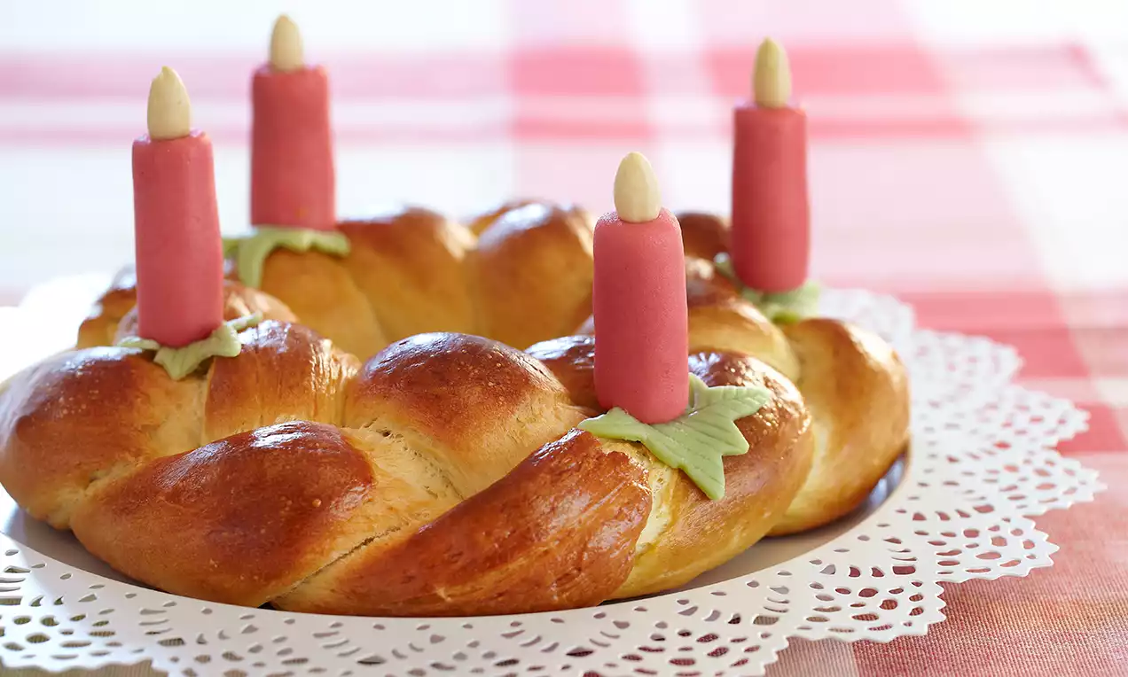 Couronne briochée dorée tressée, ornée de bougies roses avec des feuilles en pâte sur napperon ajouré et nappe à carreaux rouges