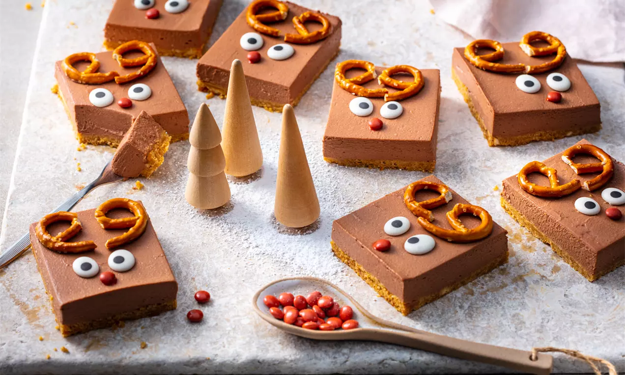 Квадратни парчета шоколадов чийзкейк върху бисквитен блат, декорирани с солени гевречета, захарни очи и червени бонбони, поставени върху светъл плот