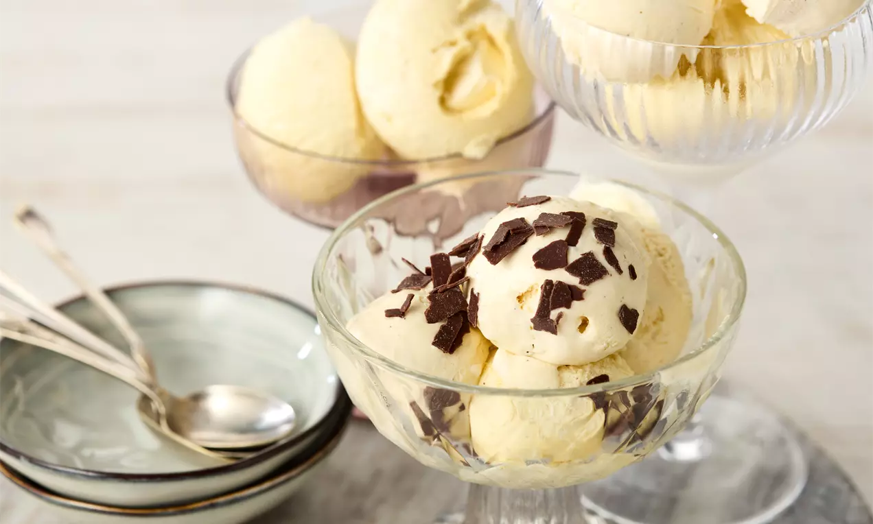 Înghețată vegana cremoasă, galben-pal, modelată în cupe din sticlă transparentă, decorată cu fulgi de ciocolată maro pe masă albă