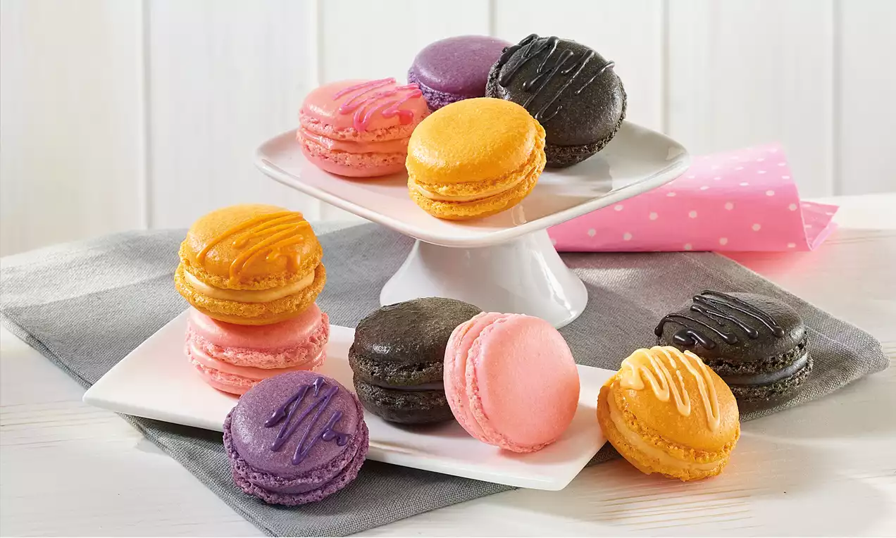 Bunte Macarons in Orange, Rosa, Lila und Schwarz mit farbiger Glasur, arrangiert auf weißen Tellern vor hellem Hintergrund und rosa Serviette