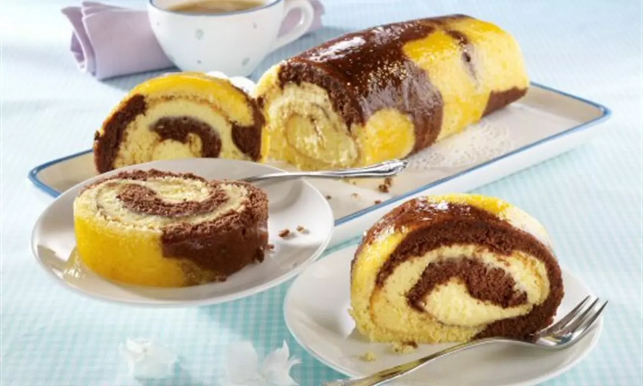 Руло с спираловидна текстура от златисто-жълт и тъмнокафяв блат, изпълнено с ванилов крем, поднесено на бял порцелан до чаша кафе