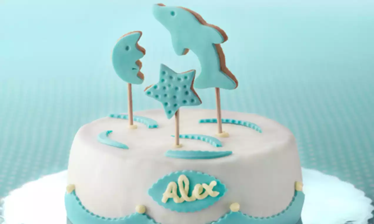 Tarta redonda cubierta de fondant blanco con detalles en azul, galletas decorativas en forma de luna, estrella y delfín sobre palillos