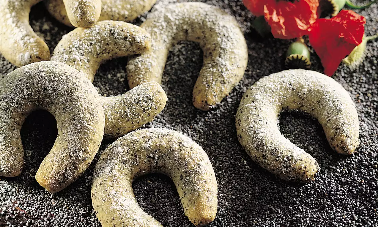 Mohnkipferl in gebogener Form, goldgelb mit dunklem Mohn bestreut, auf einem Bett aus Mohnsamen mit roten Mohnblumen dekoriert