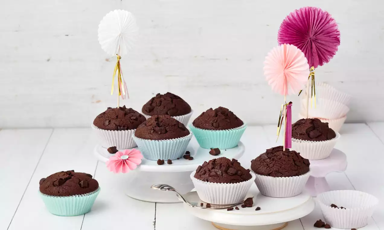Schokoladenmuffins mit dunkler, rauer Oberfläche in bunten Papierförmchen auf weißer Kuchenplatte, dekoriert mit farbigen Papierfächern