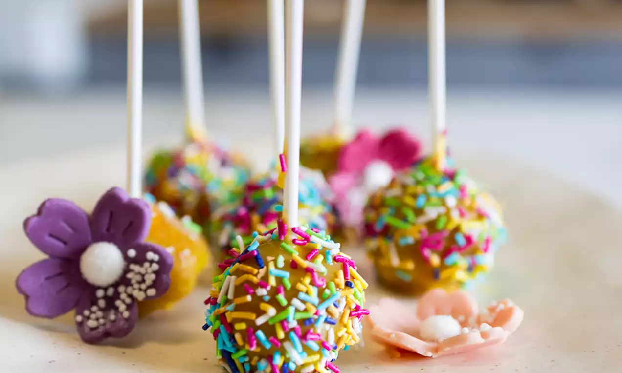 Cake popsi s bijelim štapićima, prekriveni šarenim mrvicama, na keramičkom stalku, s ukrasnim ljubičastim i ružičastim cvjetovima