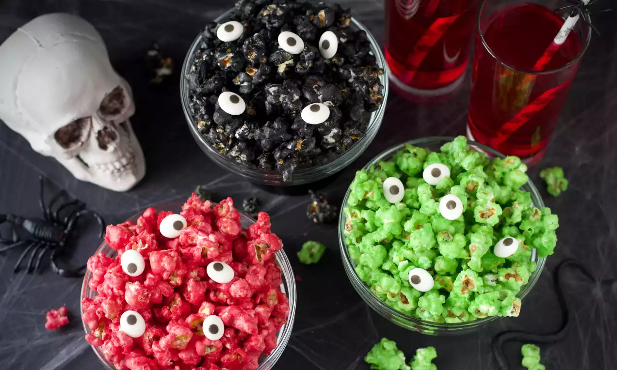 Halloween-popcorn i tre skålar, röda, svarta och gröna, med sockrig yta och vita ögon av godis, bredvid en vit dödskalle och svarta spindlar