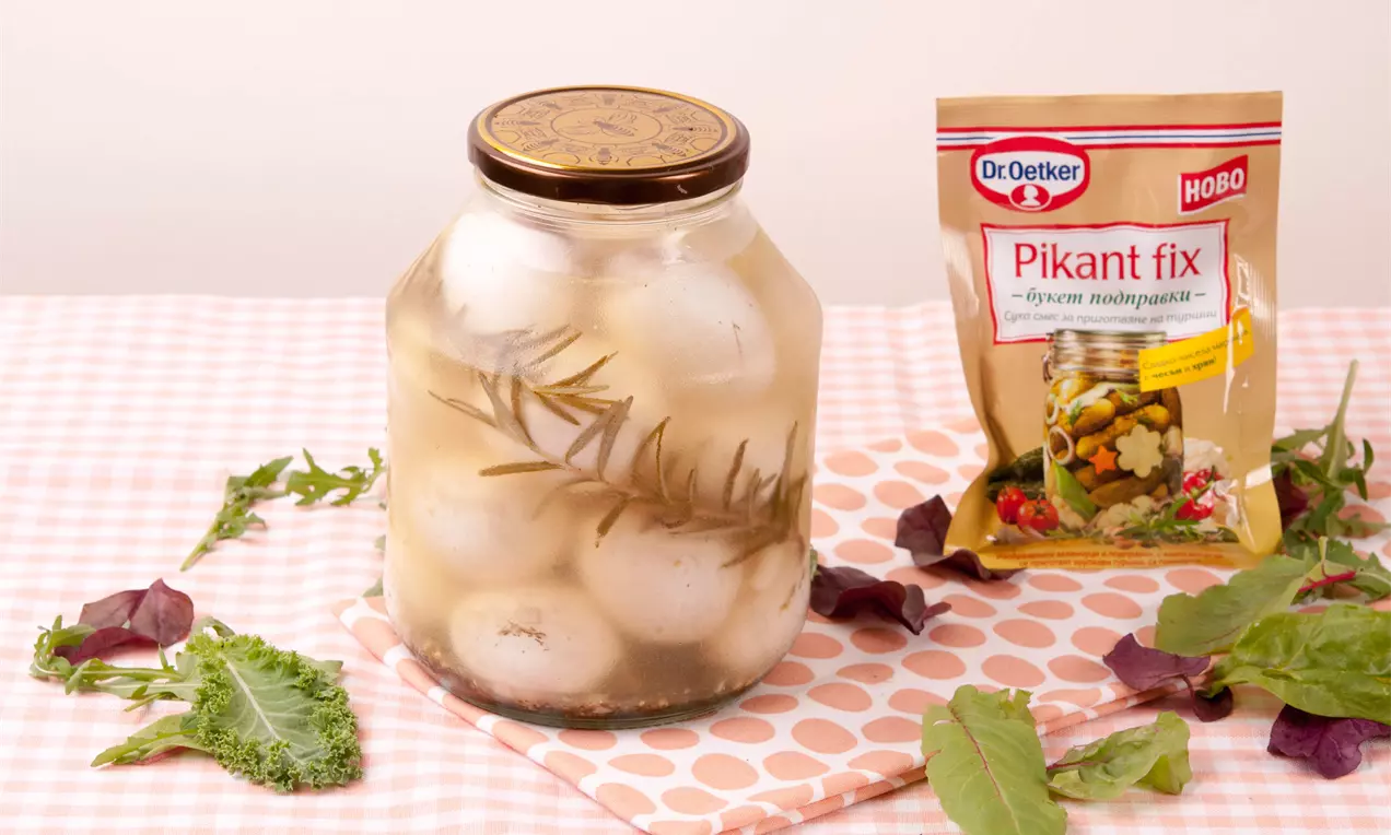 Бели мариновани яйца с гладка повърхност в прозрачен буркан, заобиколени от стръкчета розмарин и листа зеленина върху розова покривка