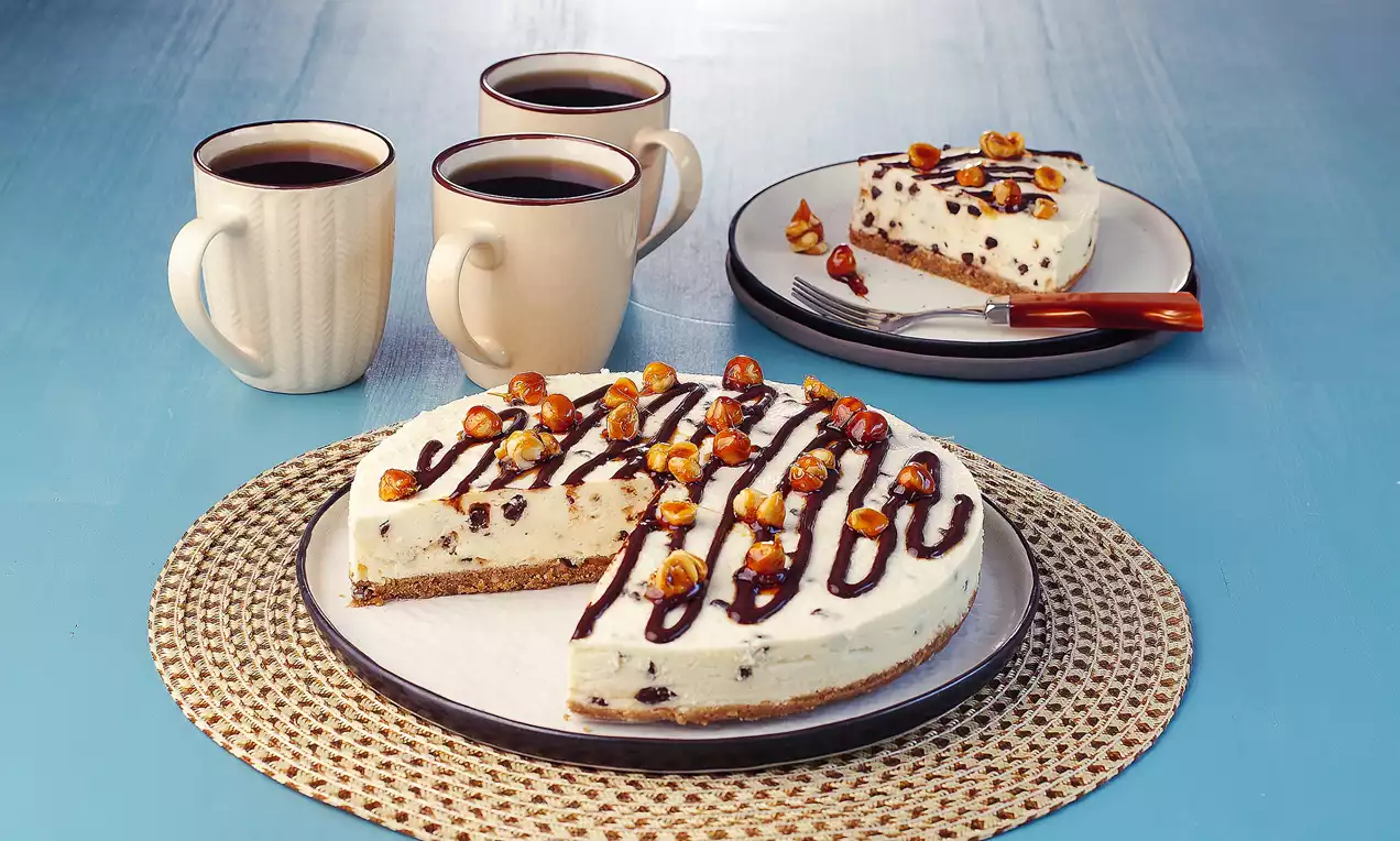 Yuvarlak bisküvi tabanlı cheesecake, beyaz kremalı iç dolgulu, üstü çikolata sosu ve fındık parçalarıyla süslenmiş, lifli bir servis altlığı üzerinde, mavi zeminde üç fincan kahve eşliğinde sunulmuş