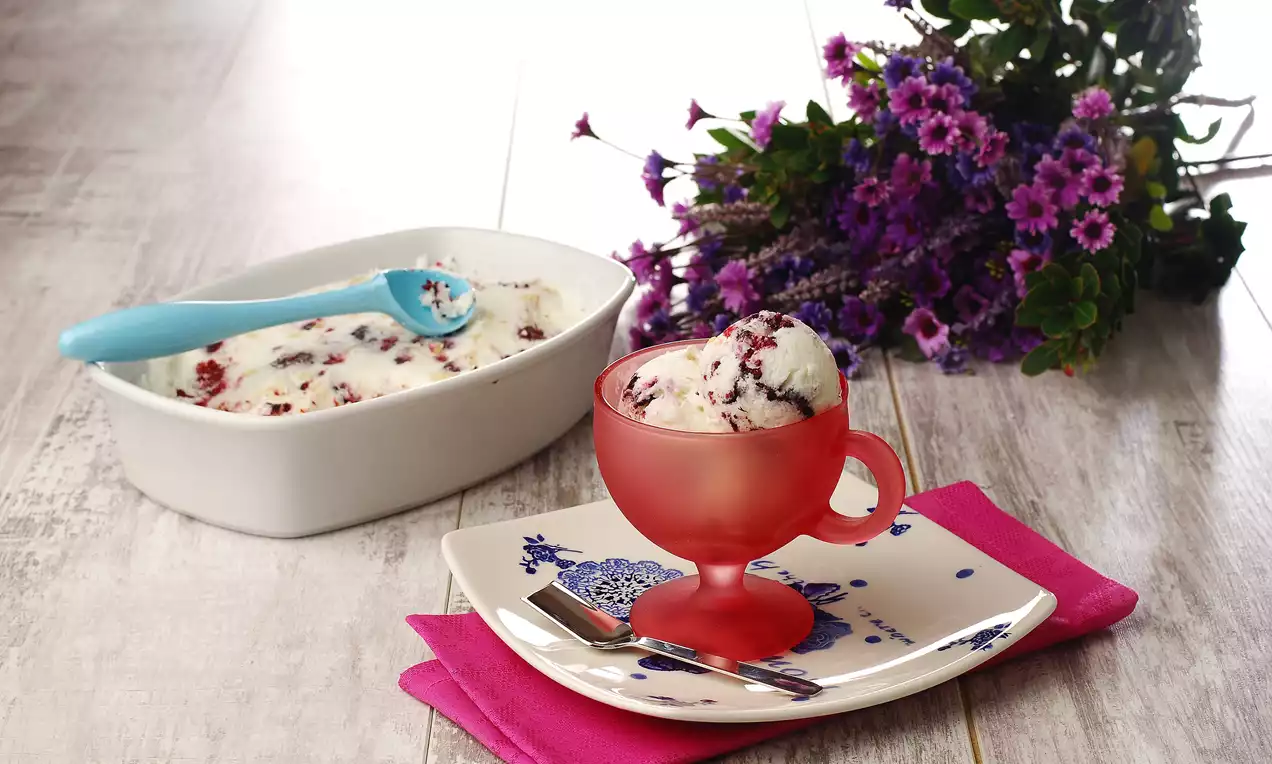 Mor çiçeklerle dekore edilmiş bir ortamda, kırmızı cam kaseye sunulmuş, mor böğürtlen parçalarıyla süslü beyaz puding