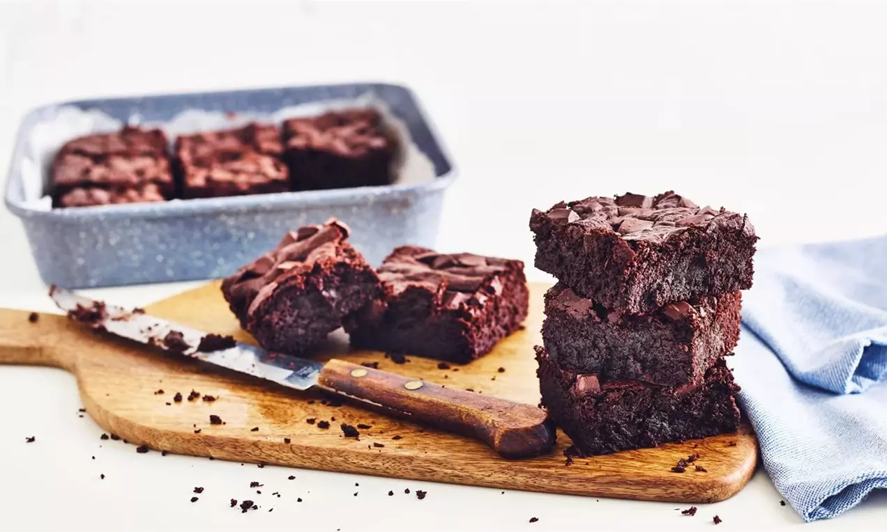 Тъмен шоколадов брауни с плътна текстура, нарязано на квадратни парчета, подредени на купчина върху дървена дъска до нож и синя кърпа
