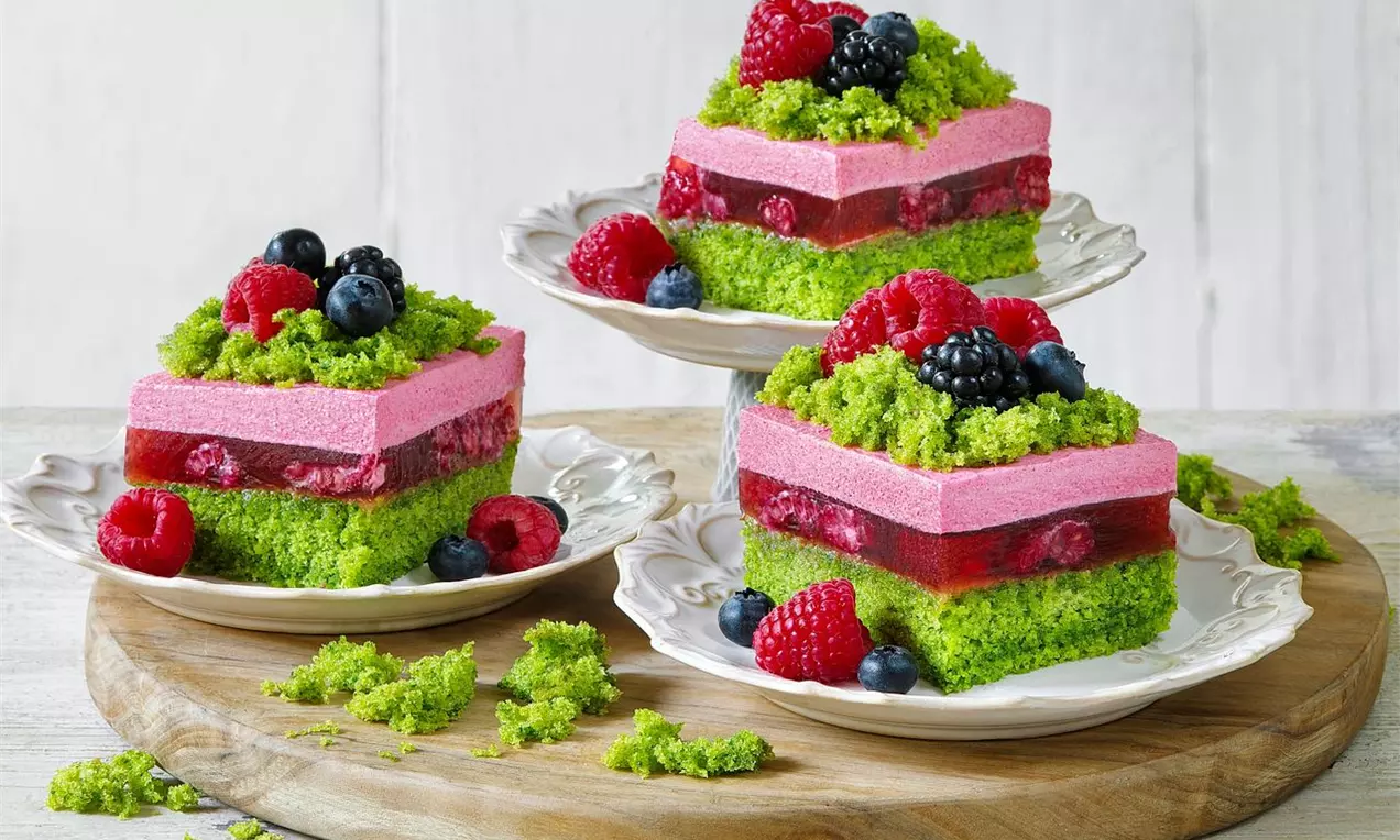 Malinové rezy s jasne zeleným piškótovým základom, krémovou ružovou vrstvou, želé s lesnými plodmi a ovocím na bielych tanieroch