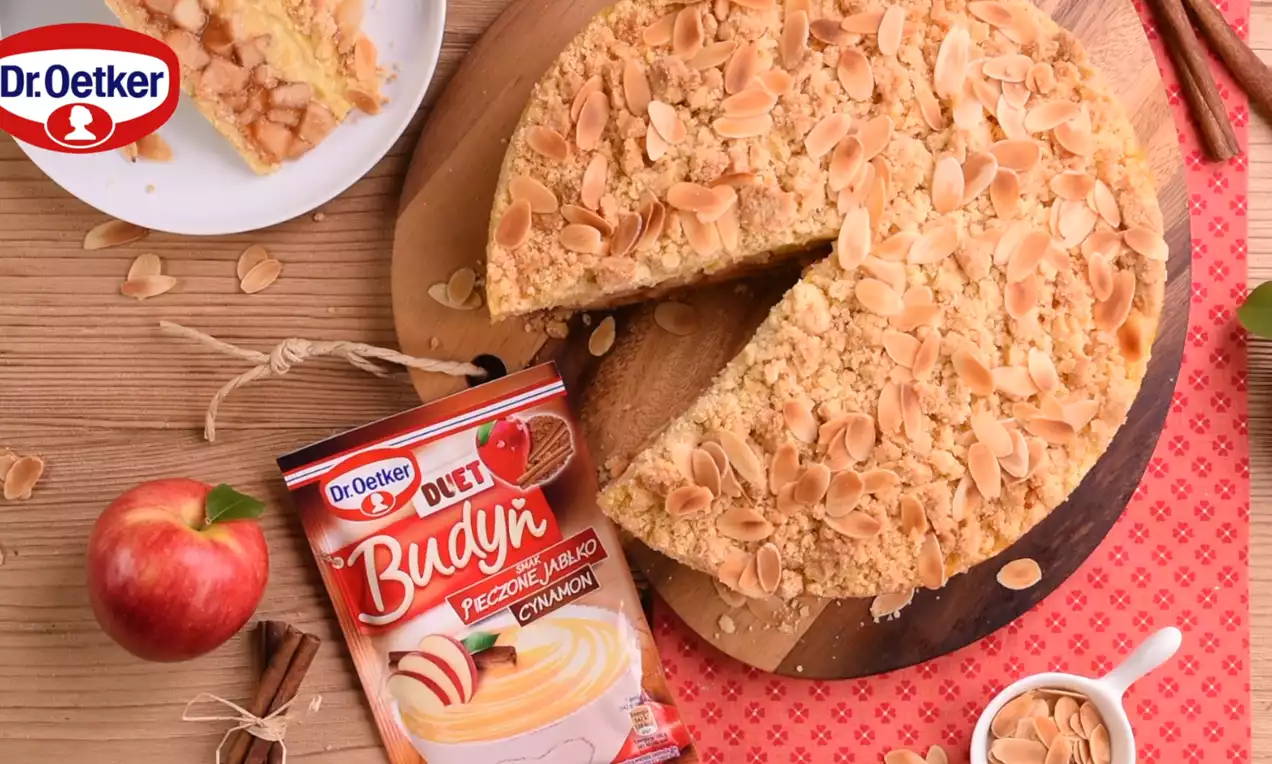 Szarlotka na drewnianym talerzu z warstwą kremowego budyniu, złotą kruszonką i prażonymi płatkami migdałów, obok jabłka i cynamon