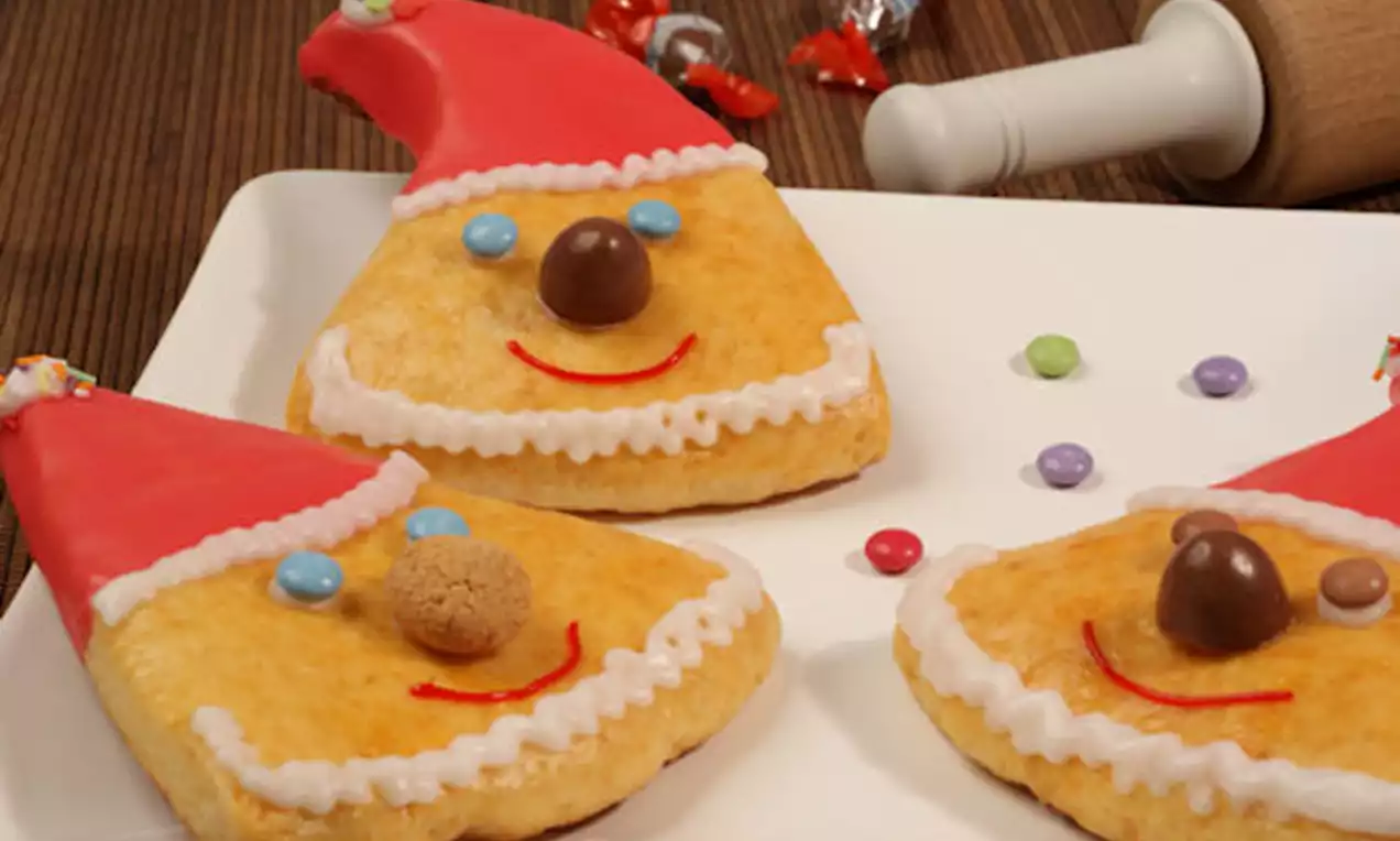 Dreieckige Nikolausgesichter aus goldenem Gebäck mit rotem Zuckermantel, Schokoladen- und Zuckerdekor auf weißer Platte angerichtet