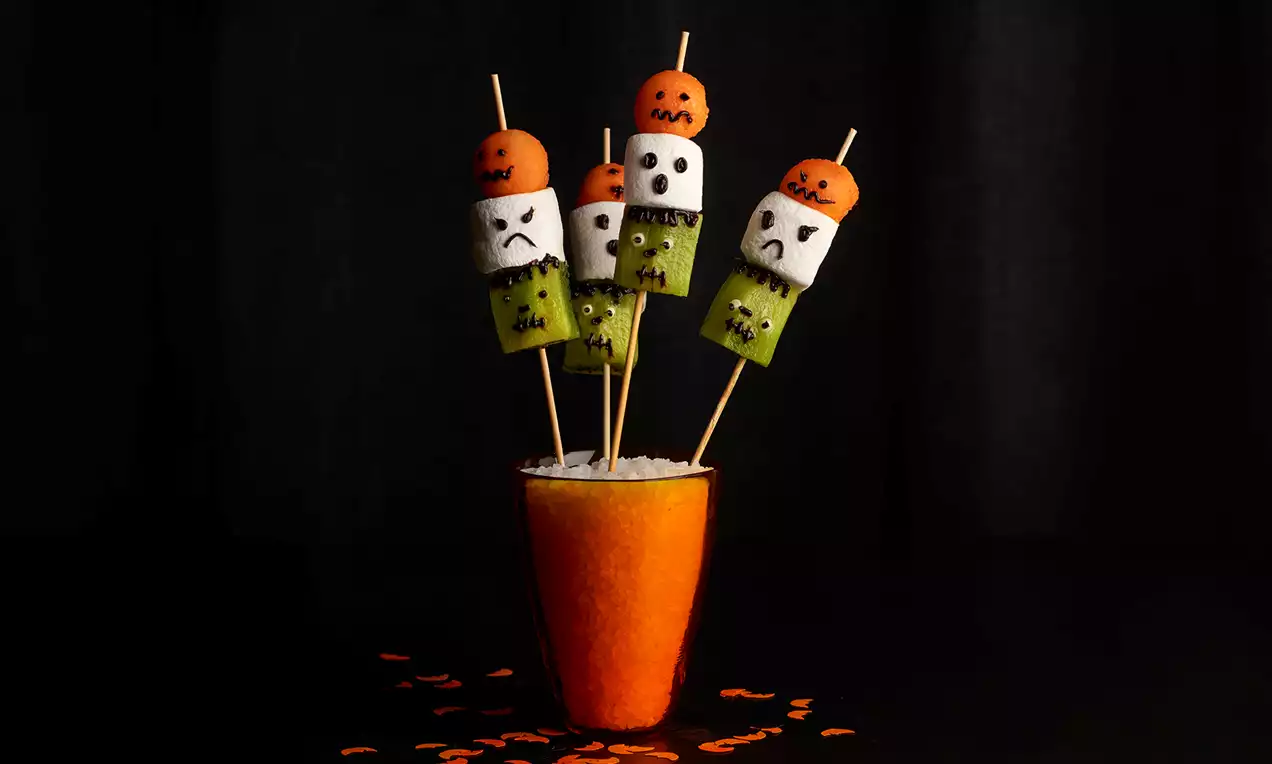 Brochetas con malvaviscos blancos pintados de caras aterradoras, trozos de kiwi verde y bolitas naranjas en palitos sobre un vaso