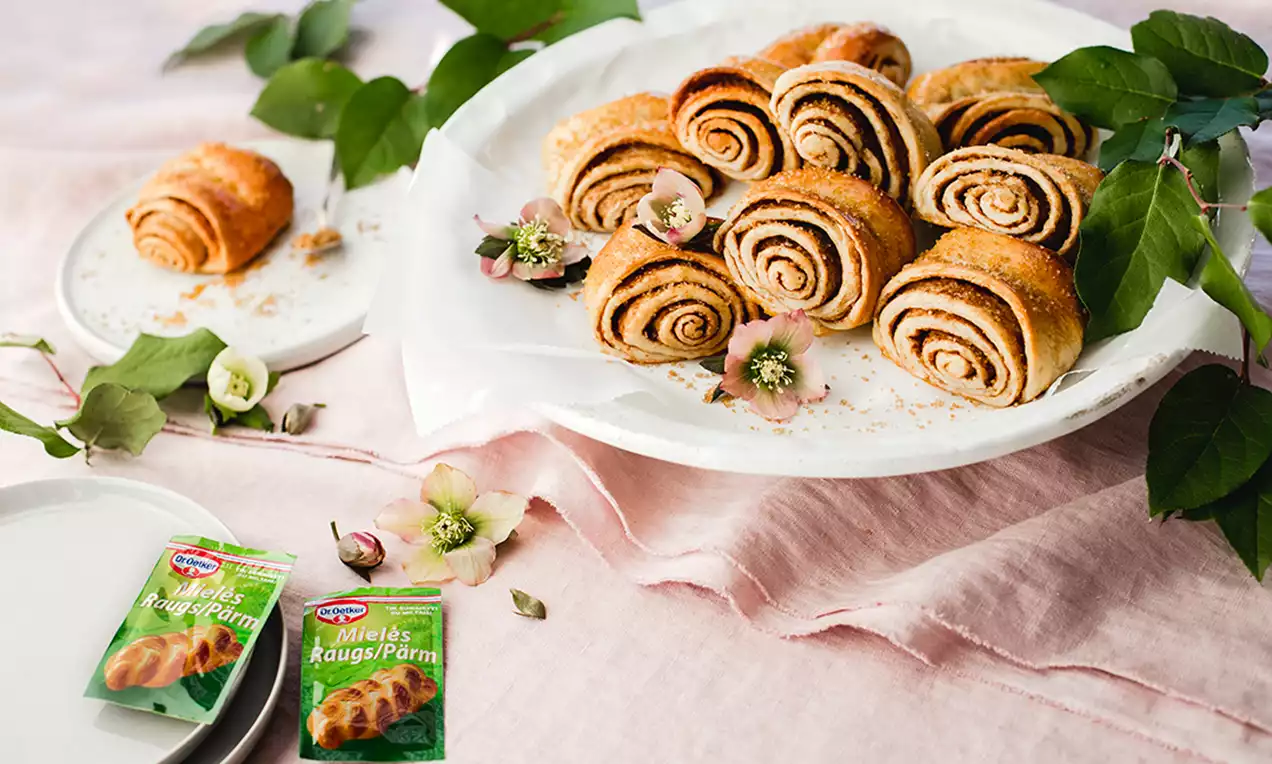 Zeltaini brūnas, spirālveida rauga maizītes ar kanēļa pildījumu uz balta šķīvja, dekorētas ar ziedošiem ziediem un zaļiem lapu zariem