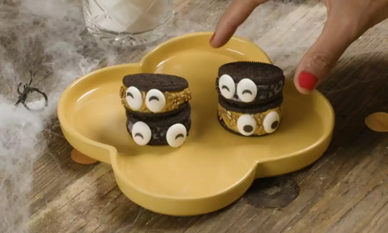 Två mörka Oreos med gul fyllning och vita ögon toppar en gul blomformad tallrik på träbord med bomullsspindelnät och en glasdetalj