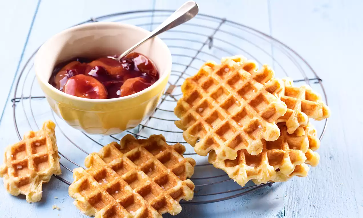 Goldgelbe Waffeln mit leichter Textur auf einem Metallrost neben einer cremefarbenen Schale mit glänzendem Pflaumen-Sauerkirsch-Kompott