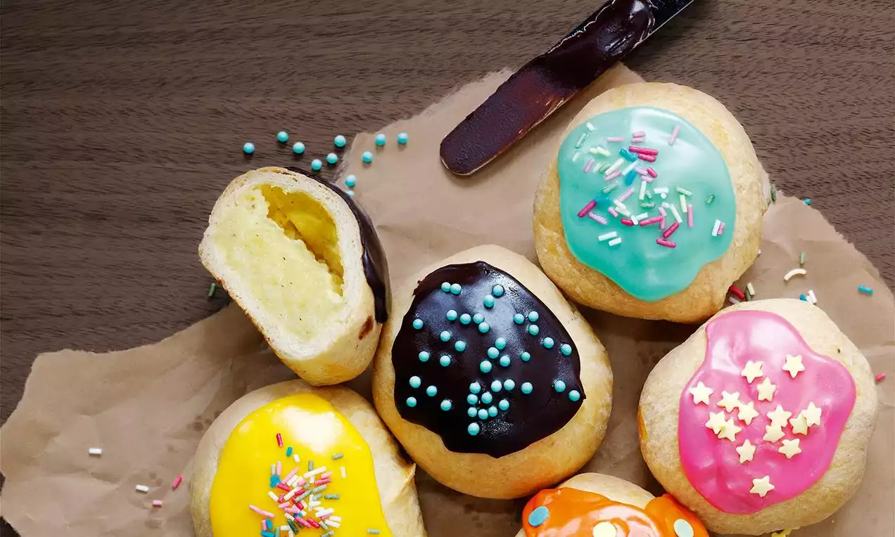 Fastelavnsboller med gul kremfyll, dekket av glasur i blått, rosa, gult, grønt og sjokolade, toppet med fargerik strøssel på matpapir