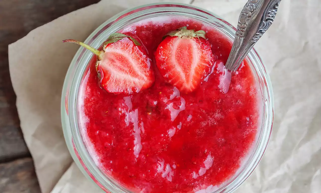 Confiture rouge brillante à la fraise avec morceaux de fruit et deux fraises fraîches sur le dessus dans un bol en verre élégant