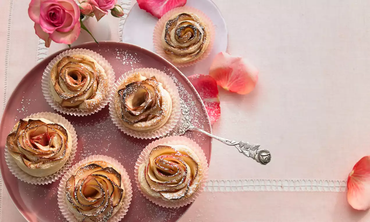 Filigranes Apfel-Rosen-Küchlein mit goldbrauner Kruste, blütenförmig drapiert, auf einem zarten Teller mit Rosenblütenblättern arrangiert