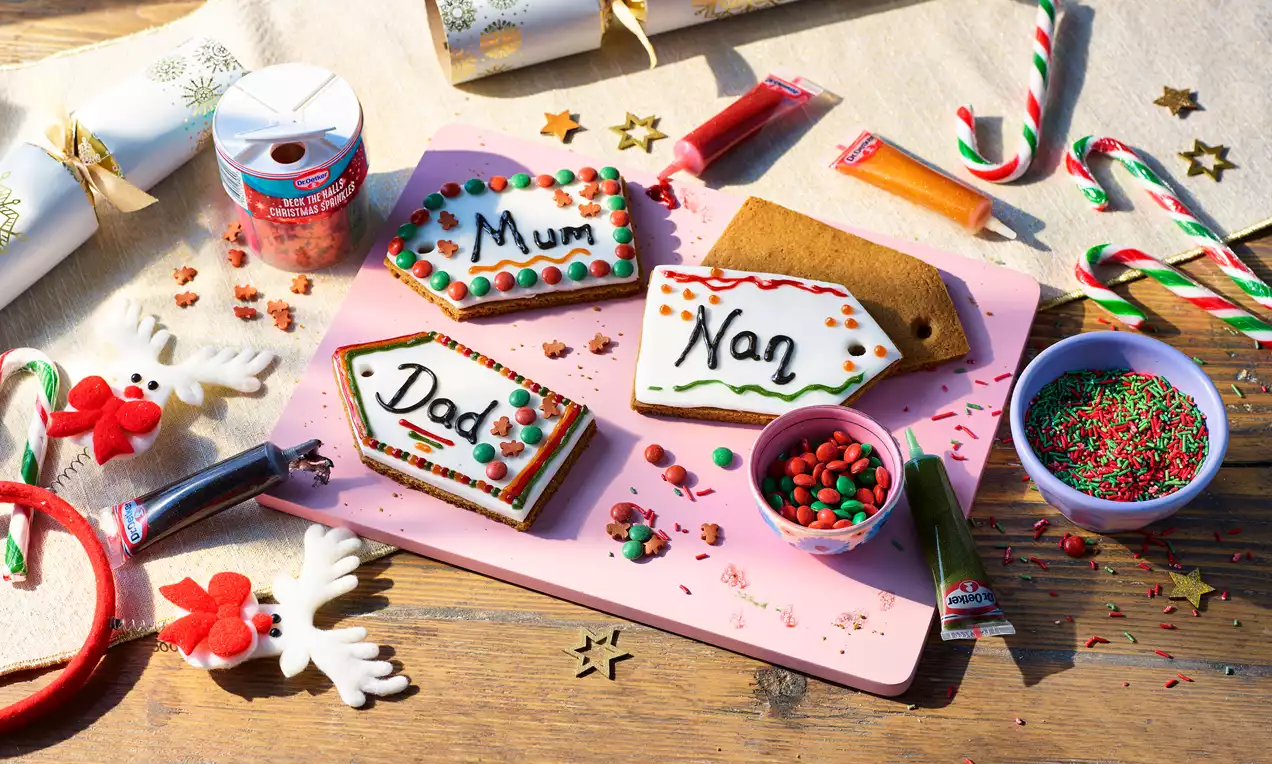 Gift tag-shaped biscuits with golden-brown surfaces, iced edges, handwritten names, candy decorations, and sprinkles on a pink board
