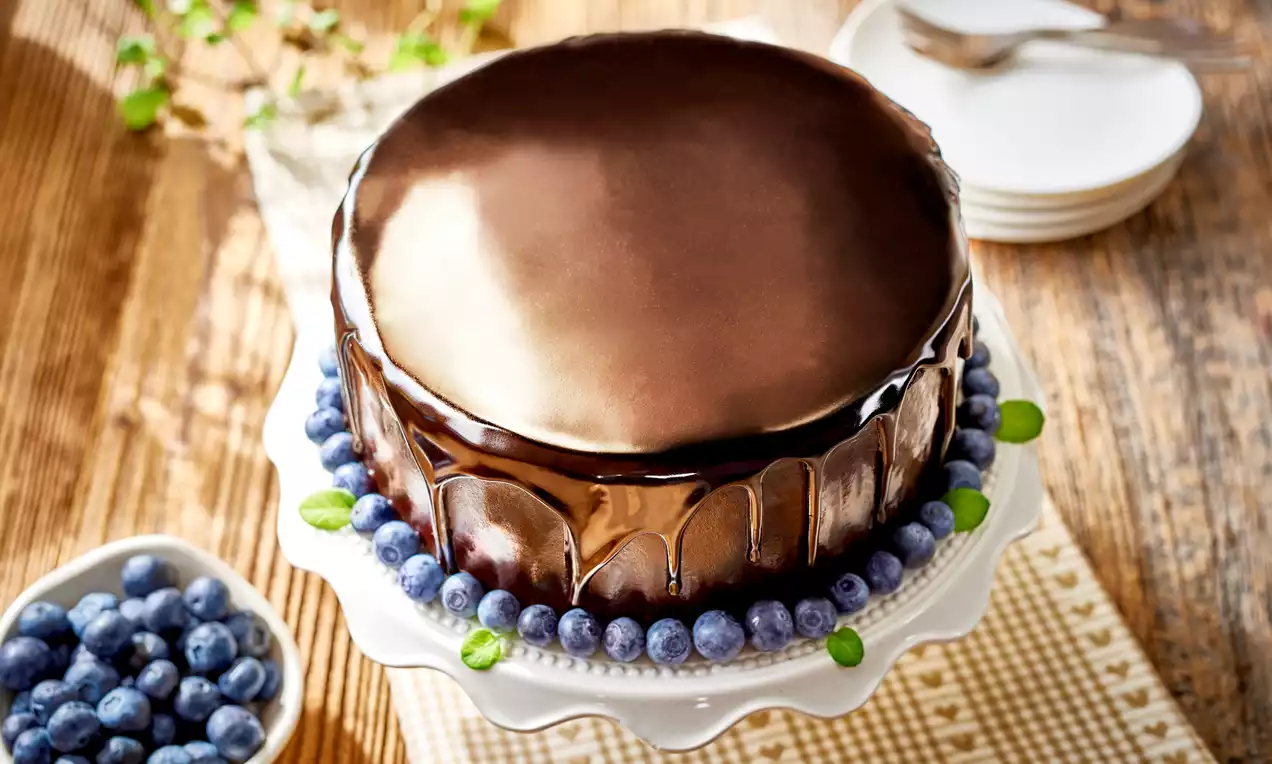 Okrągłe ciasto z gładką, lśniącą czekoladową polewą, ozdobione borówkami i listkami mięty, na białej paterze ustawionej na kratkowanym tle