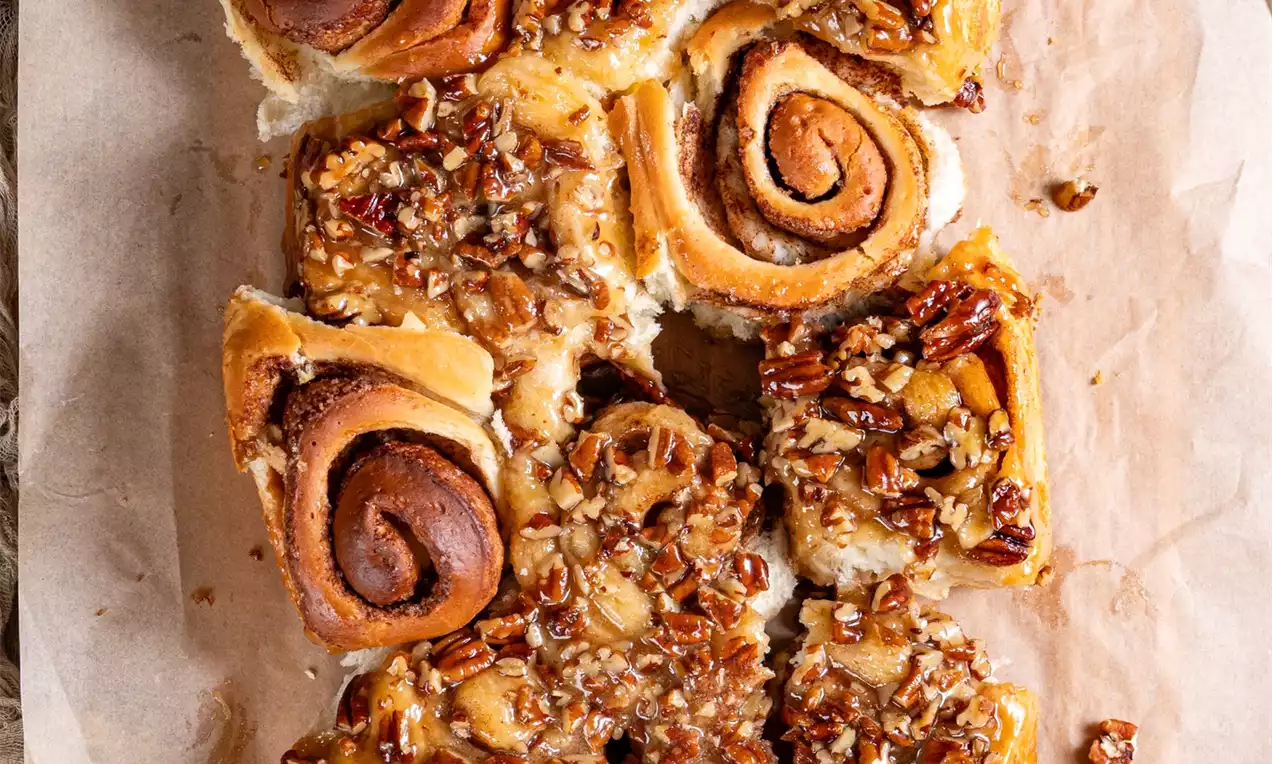 Lepljivi cimetovi polžki tople zlate barve, mehki in puhasti, prekriti s svetlečo karamelno glazuro in posuti s hrustljavimi orehi na peki papirju