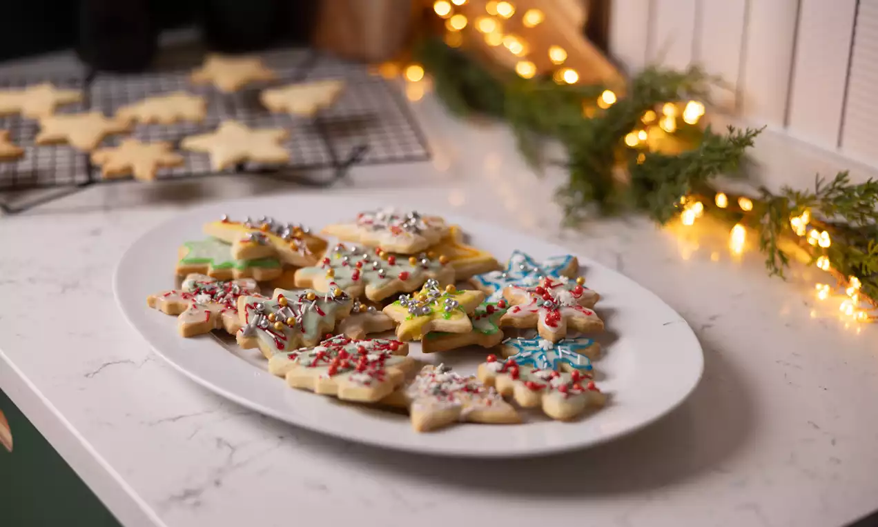 Biscuiți de Crăciun în formă de stea, glazurați în culori vibrante și presărați cu decoruri colorate, așezați pe un platou alb pe blat marmorat