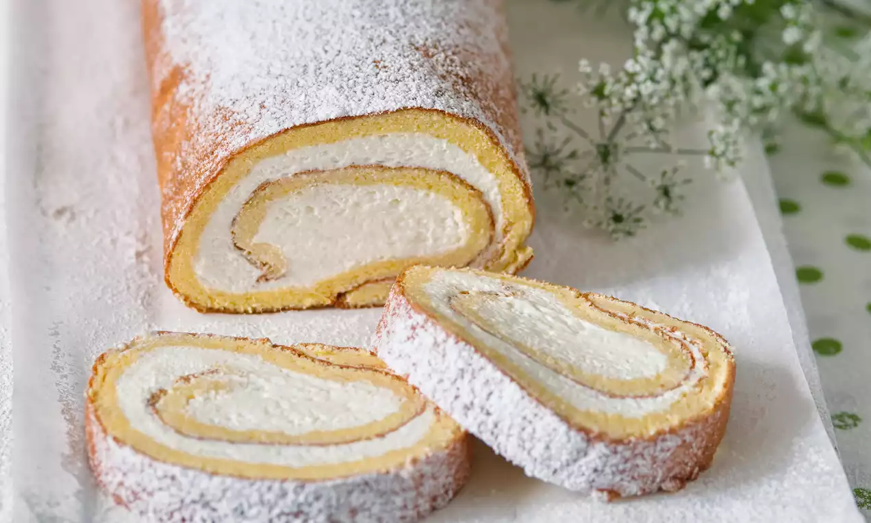 Nadýchaná, zlatavá roláda se světlým krémem, jemně pocukrovaná, na bílém podkladu; doplněná čerstvým květinovým dekorem