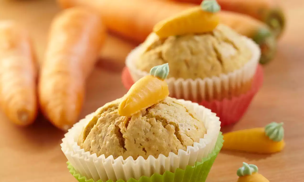 Saftige Rübli-Nuss-Muffins in bunten Papierförmchen, gekrönt mit kleinen orangefarbenen Marzipanmöhren, arrangiert auf hellem Holz mit Karotten im Hintergrund