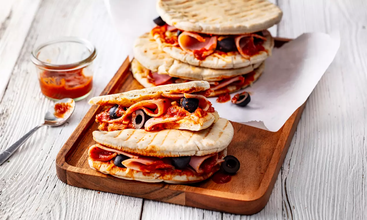 Запечени панини с тънко набръчкана коричка, пълни с доматен сос, розови резени прошуто и черни маслини върху дървена табла