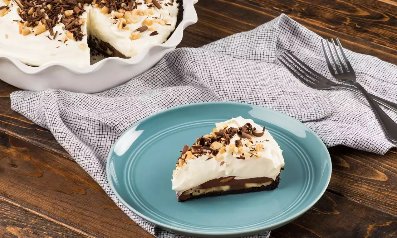 A slice of chocolate peanut butter pie with creamy white topping, chocolate shavings, and chopped nuts on a teal plate with forks nearby