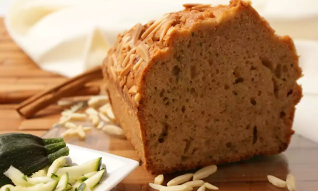 Ein goldbraunes Kuchenstück mit lockerer Textur, bedeckt von gehobelten Mandeln, umgeben von geraspelter Zucchini und Mandeln auf Holz