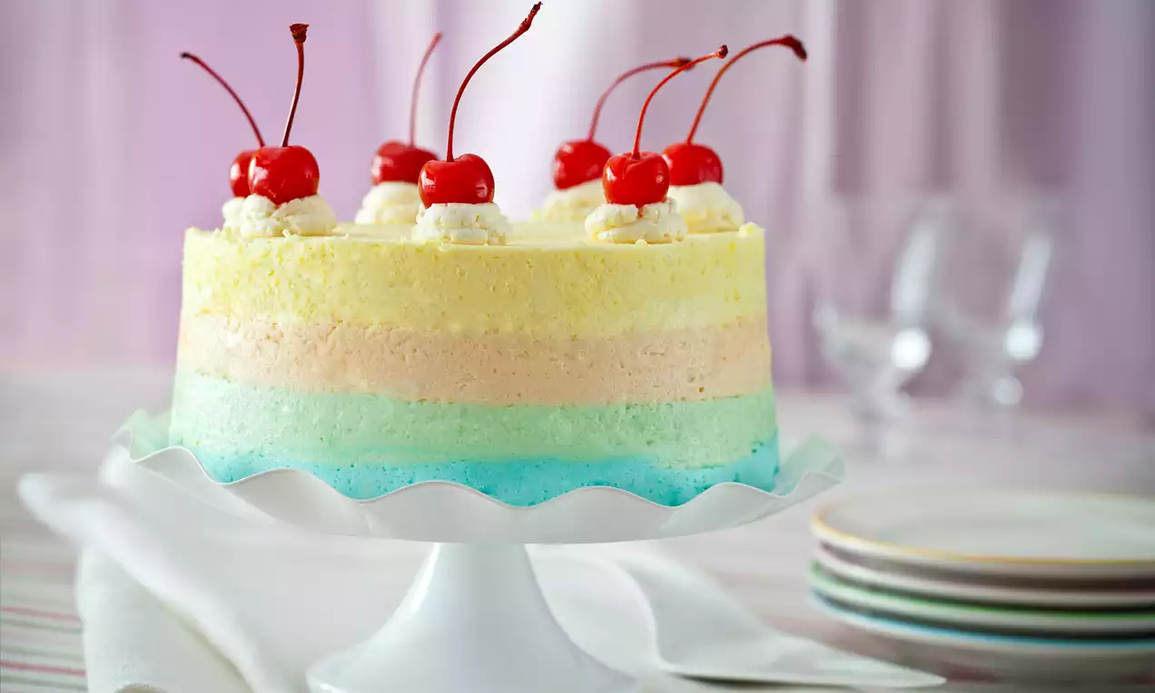 Warstwowy sernik na zimno w odcieniach tęczy, udekorowany bitą śmietaną i czerwonymi wiśniami na białej paterze z falowanym brzegiem