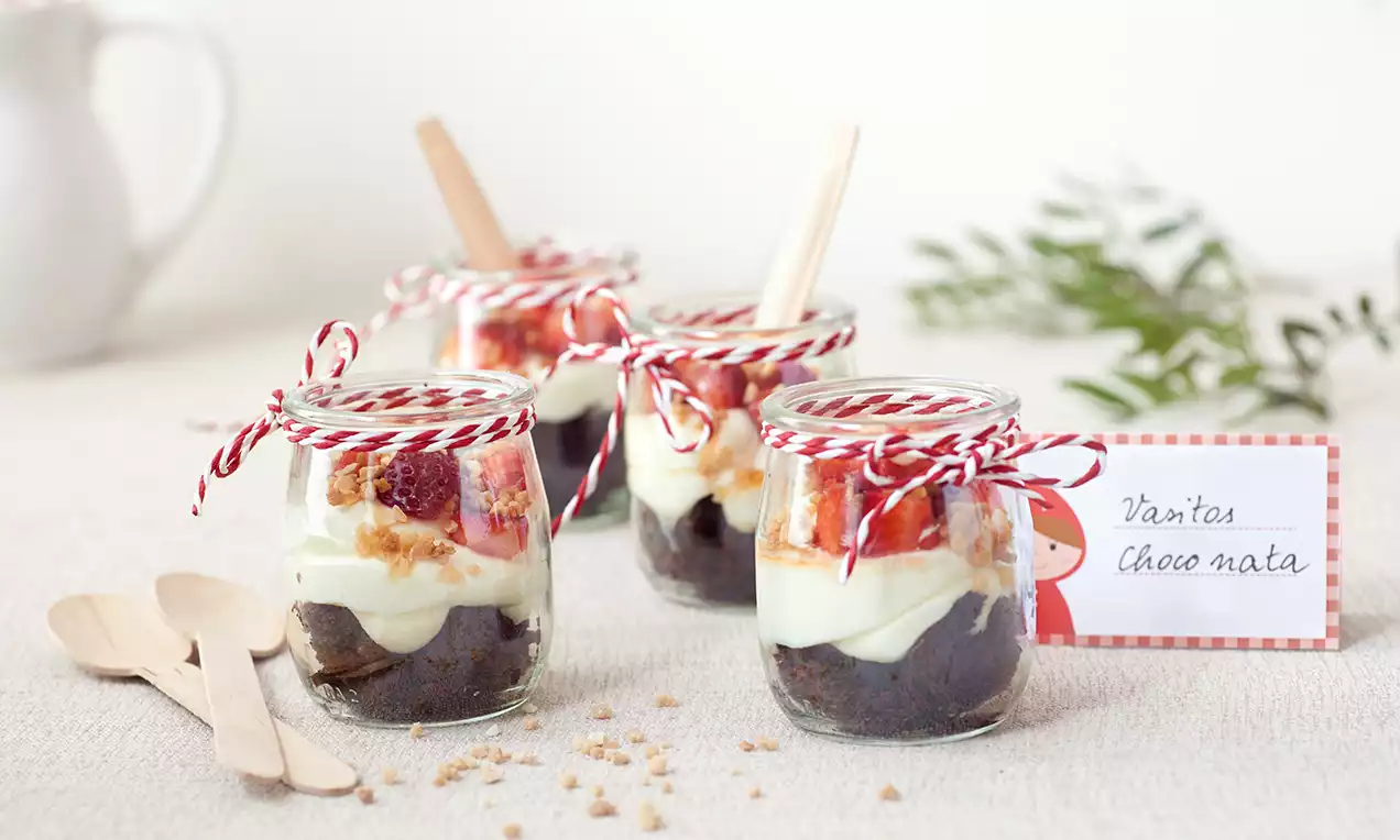 Vasitos transparentes con capas de bizcocho oscuro, crema blanca suave, trozos de fresa y decoración con lazos rojos sobre una mesa clara