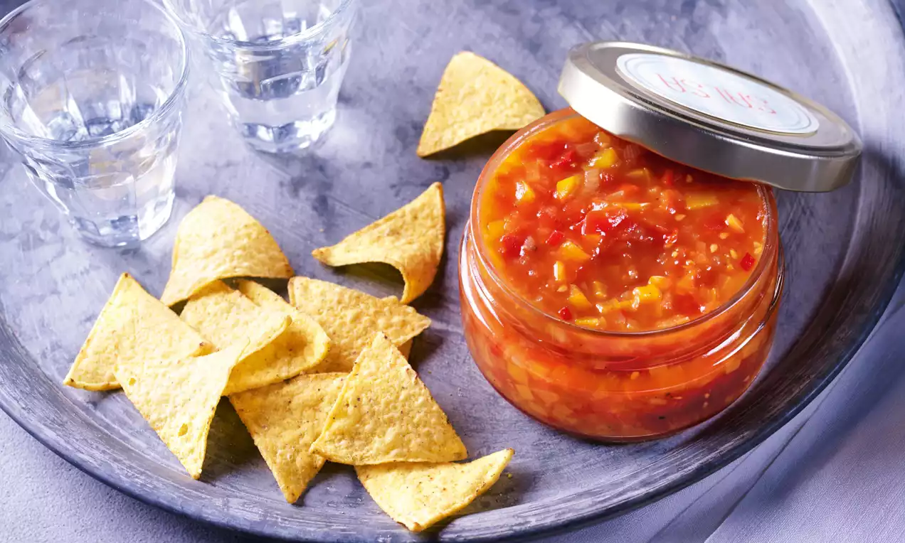 Salsa rouge orangée aux morceaux de tomate et poivron dans un bocal en verre, entourée de chips dorées sur un plateau gris