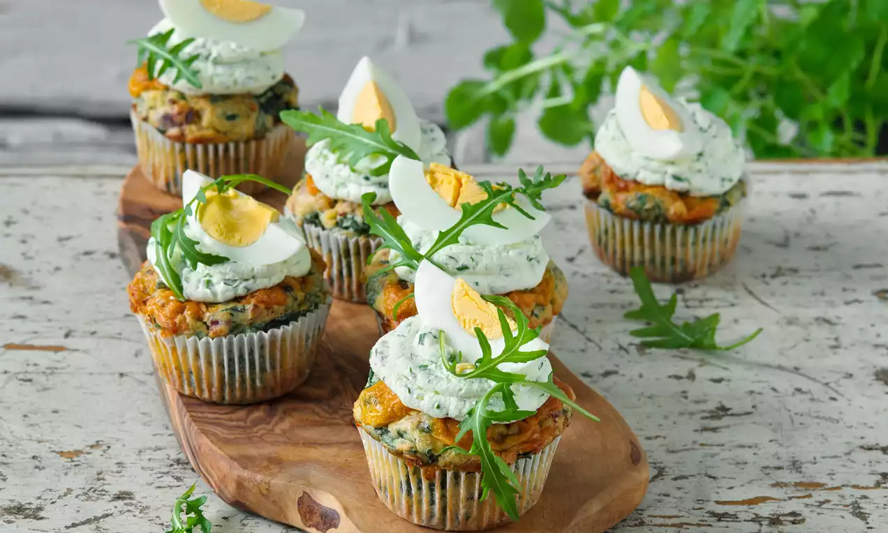 Velikonoční slané muffiny s křupavou zlatohnědou texturou, vrstvou zeleného bylinkového krému, plátky vajec a lístky rukoly na dřevěném prkénku