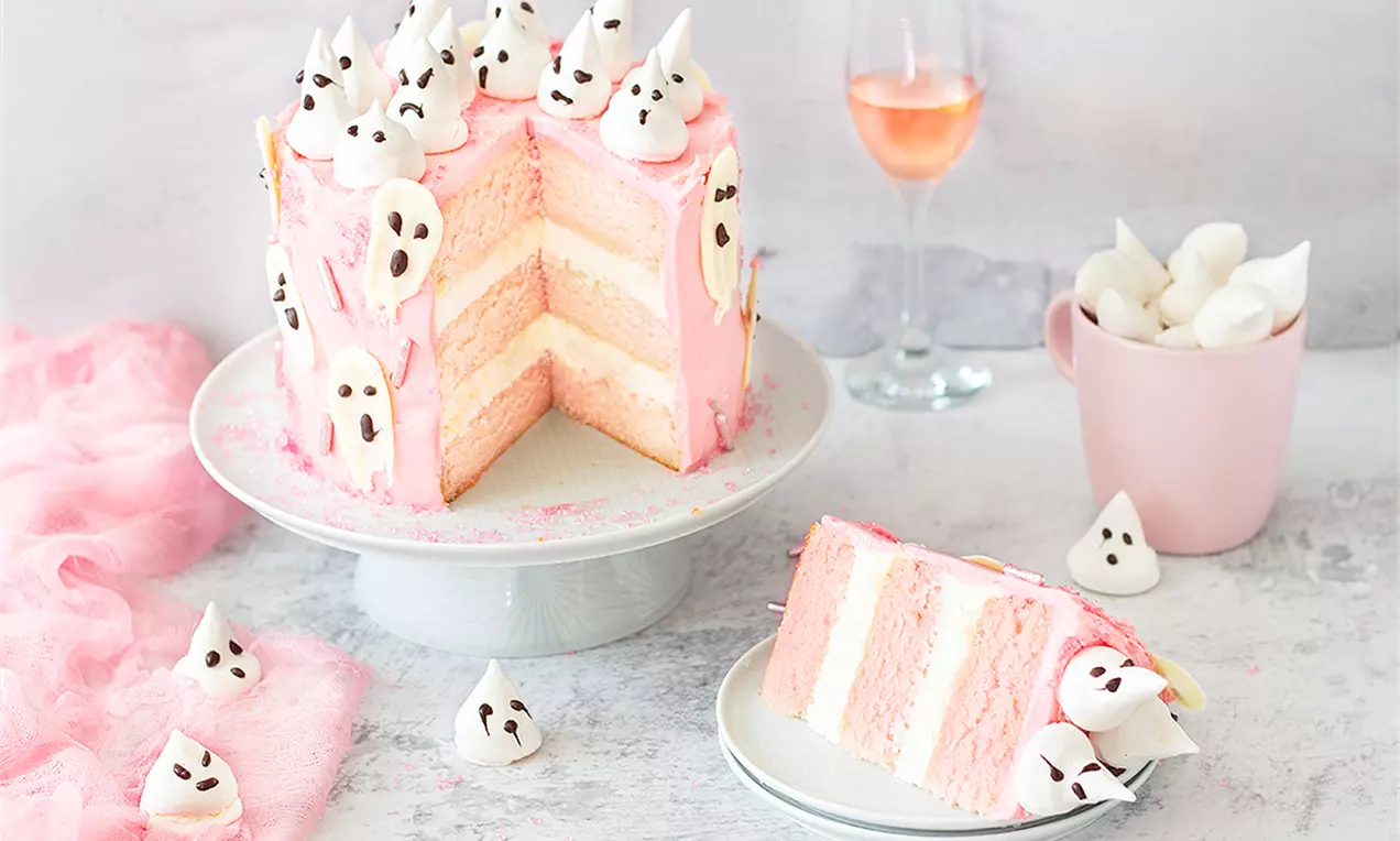 Висока розова торта, украсена с малки бели призрачни фигурки, с резен, разкриващ пластове крем и блат върху бяла чиния