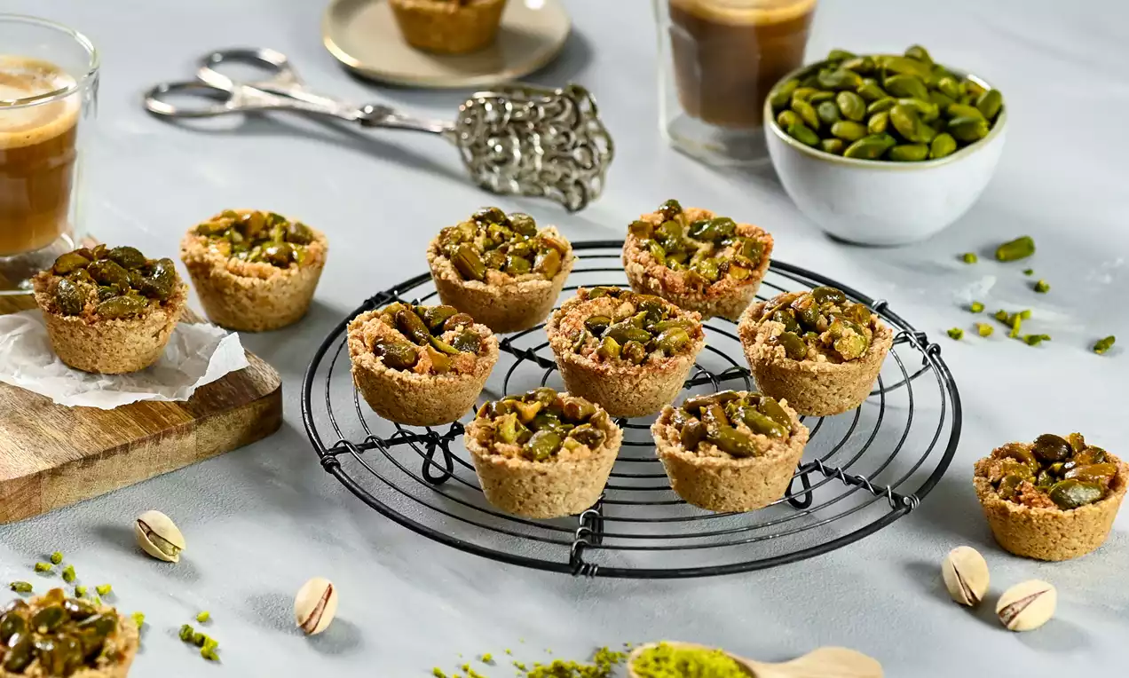 Fındık tabanlı mini granola tartlar, kıyılmış yeşil Antep fıstıklarıyla dolu, tel ızgarada sunulmuş ve etrafı fıstıkla dekore edilmiş