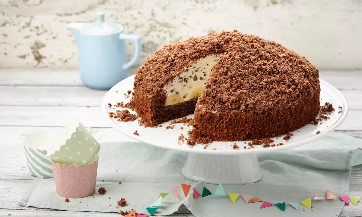 Krtičnjak kolač s tamnosmeđom čokoladnom podlogom i kremastim svijetlim punjenjem, pokriven mrvicama, na bijelom stalku okružen dekoracijom