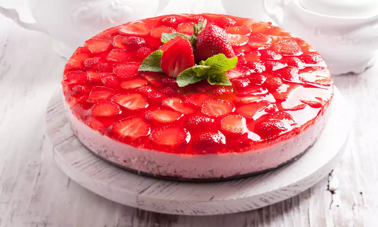 Prăjitură rotundă cu cremă roz de smântână, acoperită cu felii de căpșuni strălucitoare și decorată central cu frunze verzi și căpșuni întregi