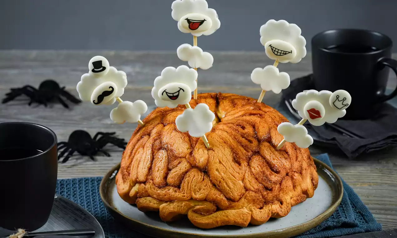 Kuchen in Form eines Gehirns mit geriffelter Oberfläche in warmem Orange auf einem Teller, verziert mit weißen Wolken-Pieksern mit Gesichtern