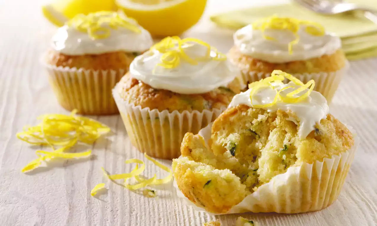 Golden cupcakes with flecks of green courgette, topped with creamy white frosting and bright yellow lemon zest on a wooden surface