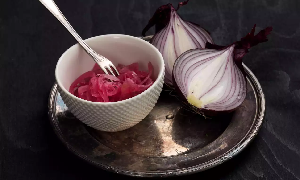 Picklad rödlök i ceriserosa strimlor i vit skål med sked, bredvid halverad rödlök på mörk metallbricka mot svart bakgrund