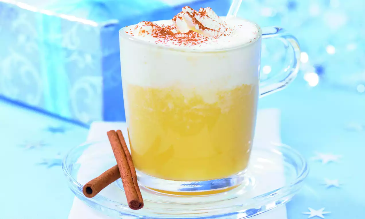 Weißwein-Orangen-Punsch in einer gläsernen Tasse mit cremigem Milchschaum, Zimtstreu und Zimtstangen auf hellem Untergrund serviert