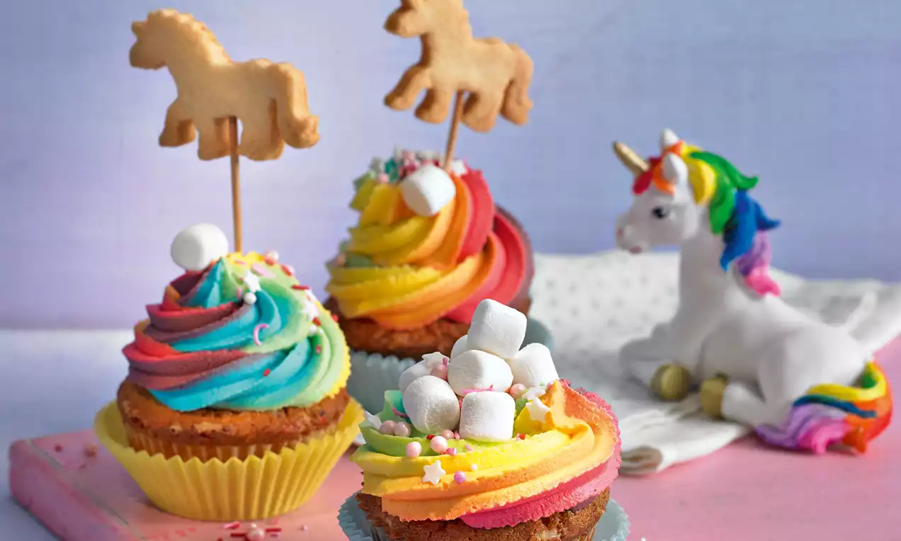 Regenbogen-Cupcakes mit buntem, spiralförmigem Frosting, kleinen Marshmallows und Keks-Einhörnern, dekoriert auf Pastell-Untergrund