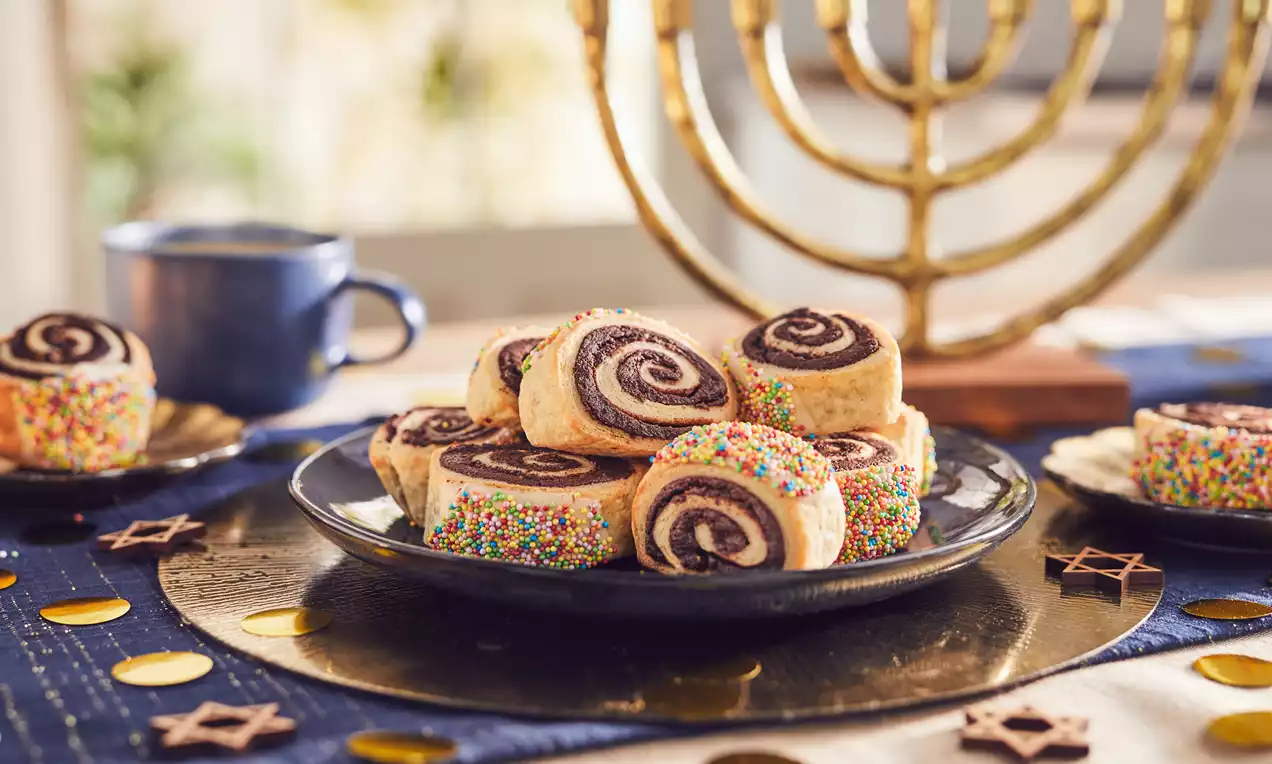 Golden spiral-shaped rugelach with a chocolate filling, some edges coated in colourful sprinkles, arranged on a dark plate